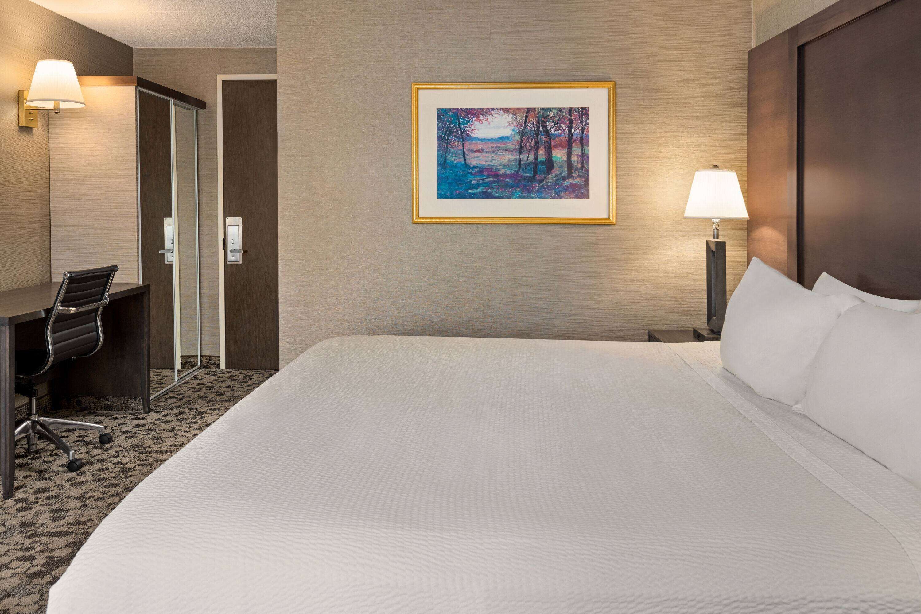 Habitación de hotel con una cama grande, un escritorio con silla, un cuadro en la pared y dos lámparas de noche.