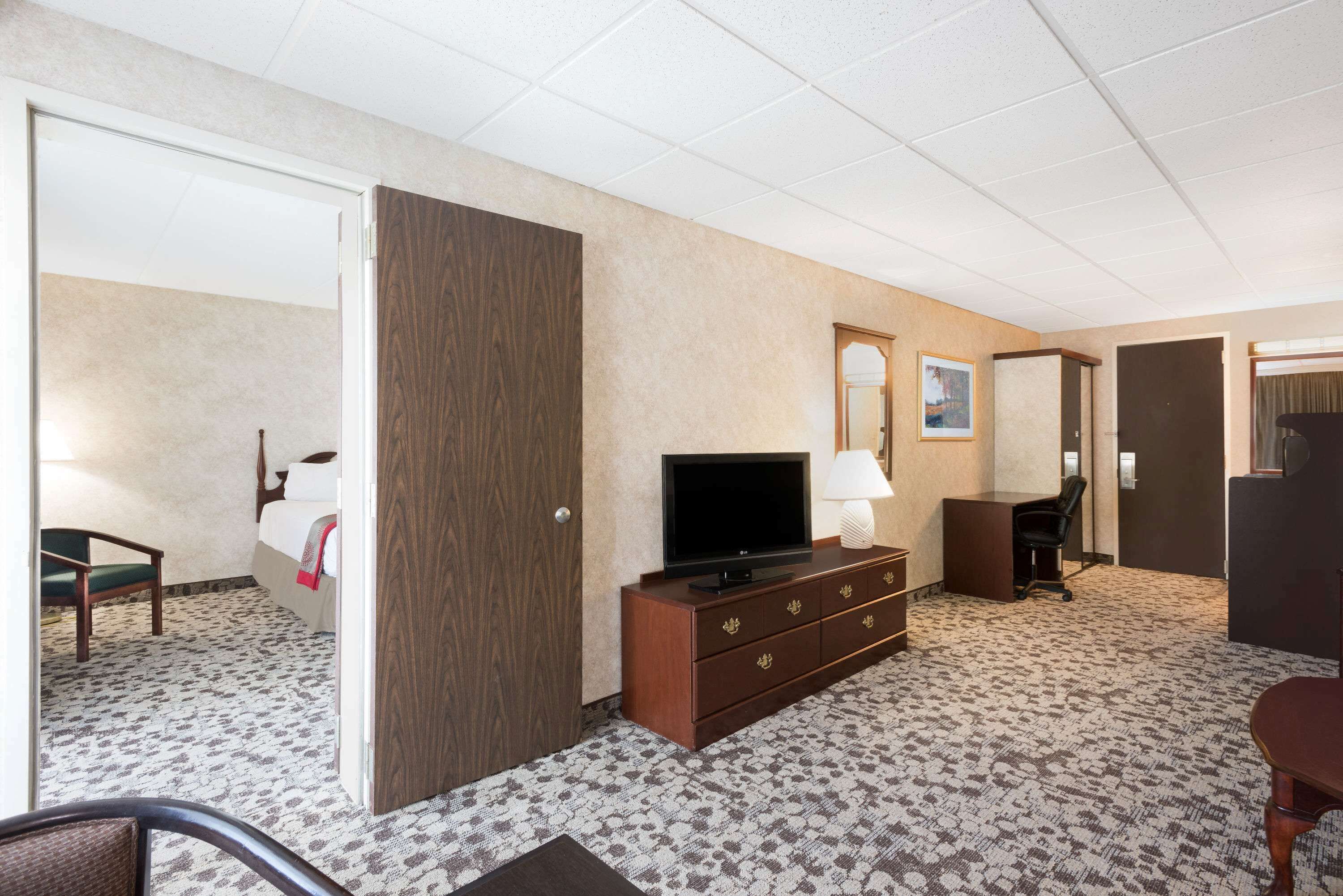 A hotel room with a bed, a TV on a wooden dresser, a desk with a chair, and a door leading to another room.