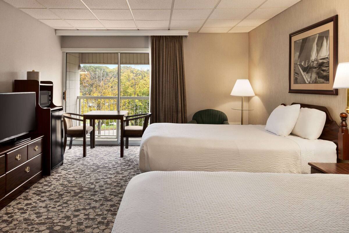 A hotel room with two beds, a desk with a chair, a television, and a window with a view of trees.