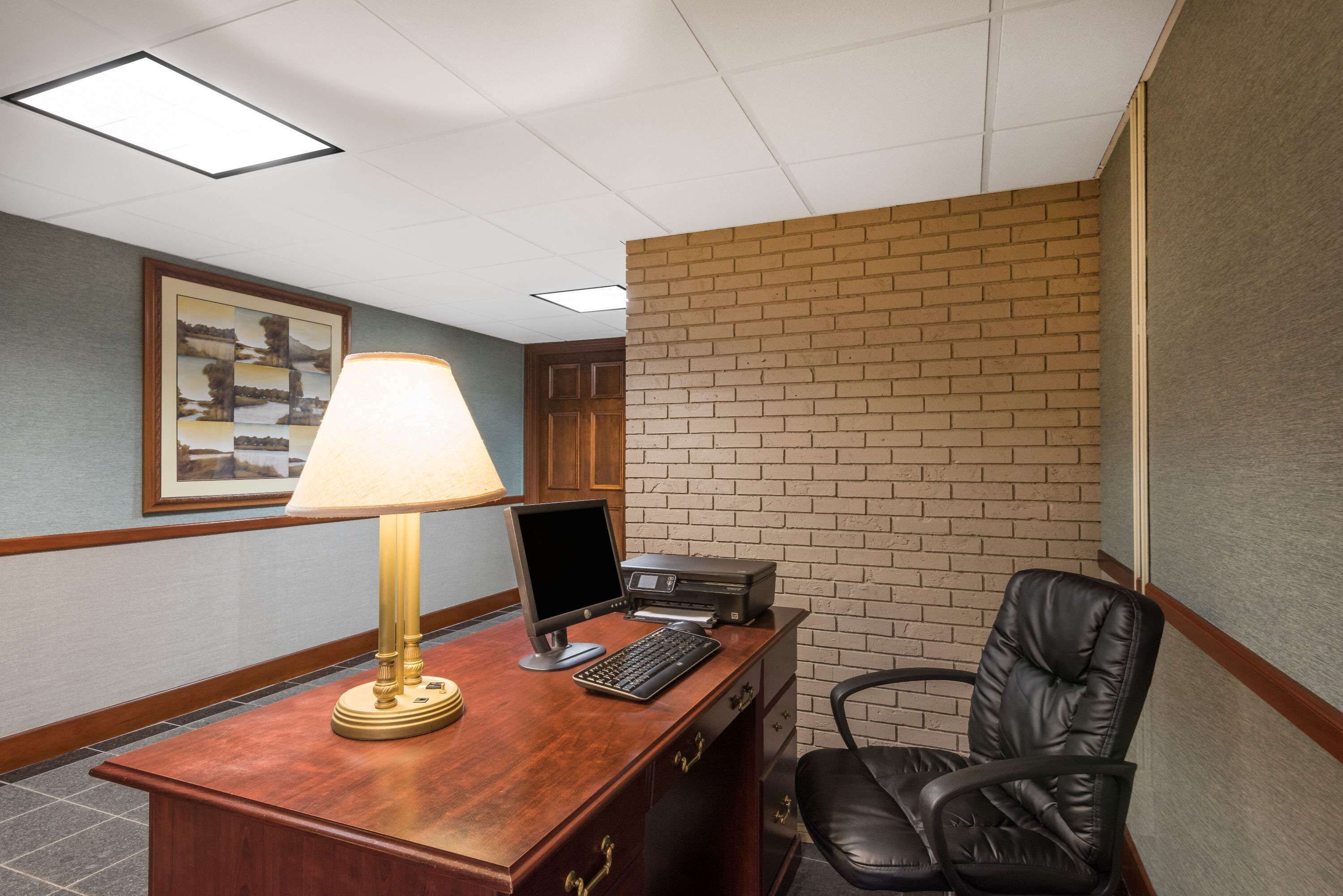 A hotel room with a wooden desk, a computer, a chair, a lamp, and a framed picture on the wall.