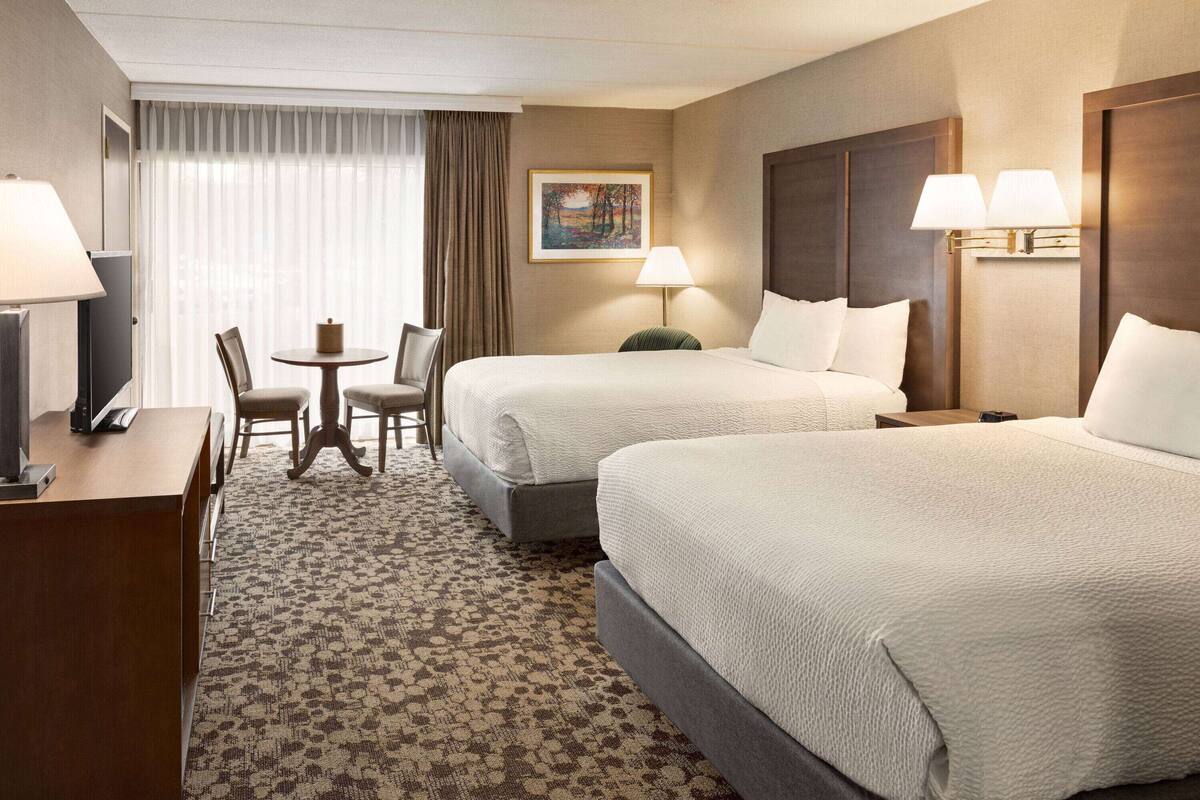 A hotel room with two beds, a desk, a chair, a lamp, and a painting on the wall.