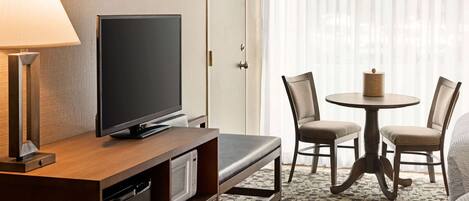 A hotel room with a flat-screen TV on a wooden desk, a lamp, a small table with two chairs, and a patterned carpet.