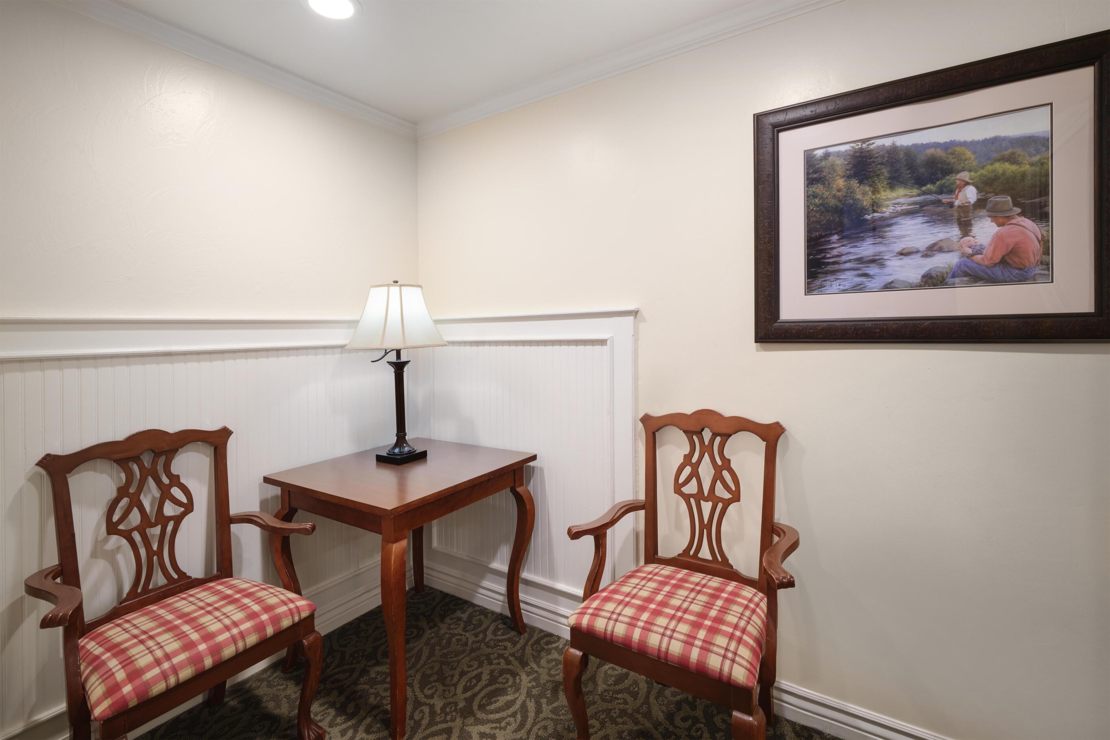 A corner room with two wooden chairs with plaid cushions, a small table with a lamp, and a framed picture on the wall.