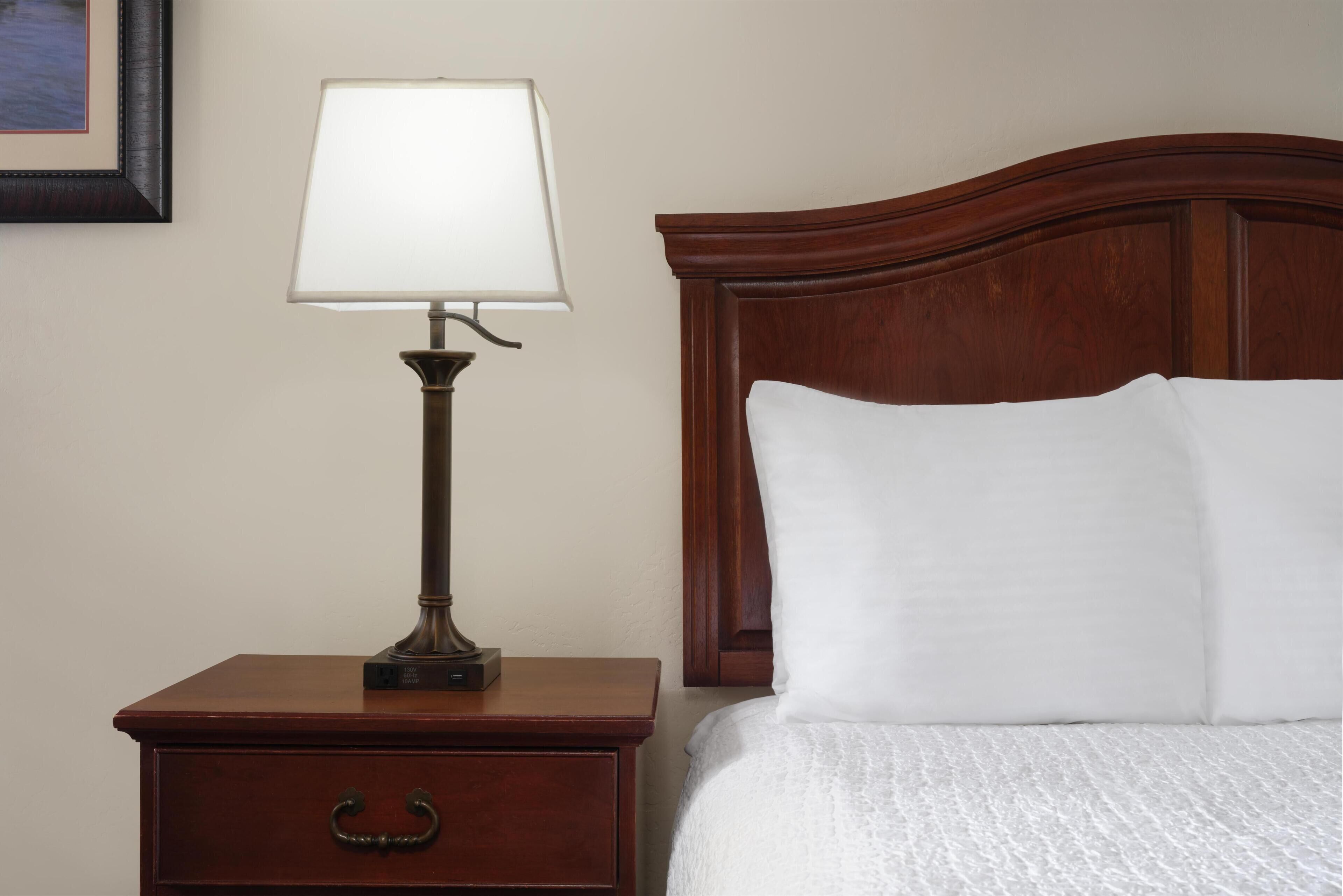 A wooden nightstand with a lamp and a drawer, a bed with white bedding, and a wooden headboard.