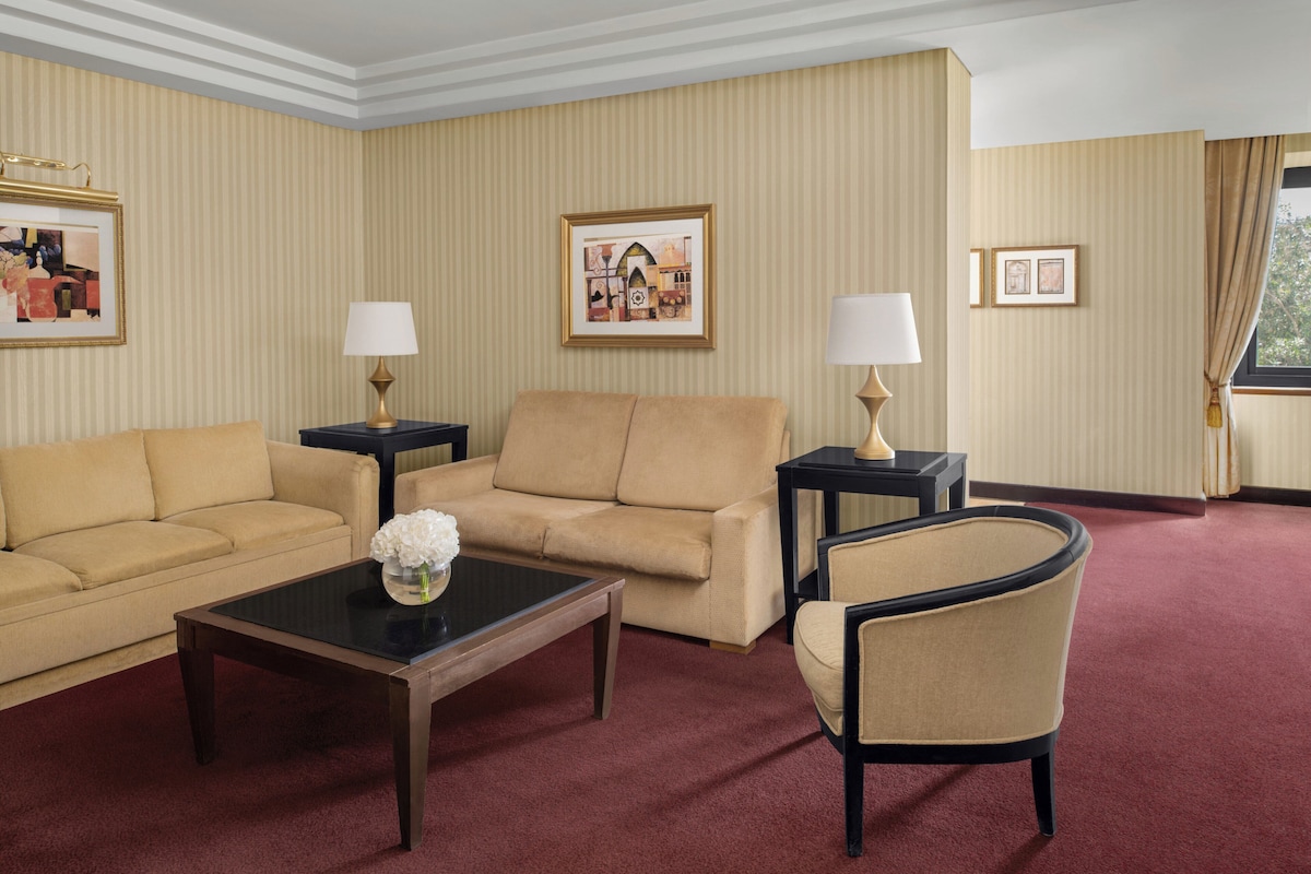 Habitación de hotel con un sofá color beige, un sillón a juego, una mesita con un jarrón de flores, dos mesitas de noche con lámparas y cuadros en las paredes.