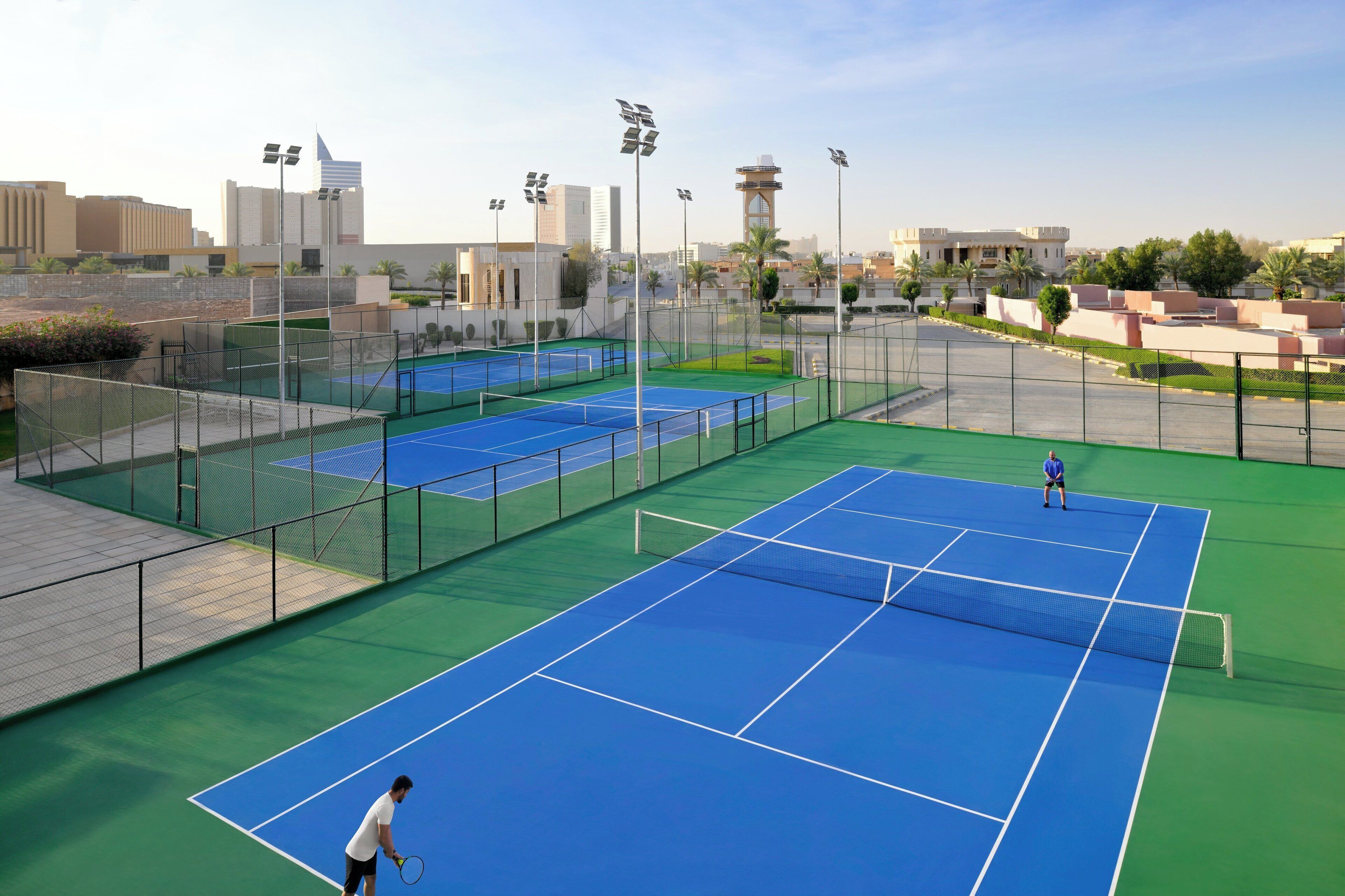 A tennis court with two players, surrounded by a fence and palm trees.