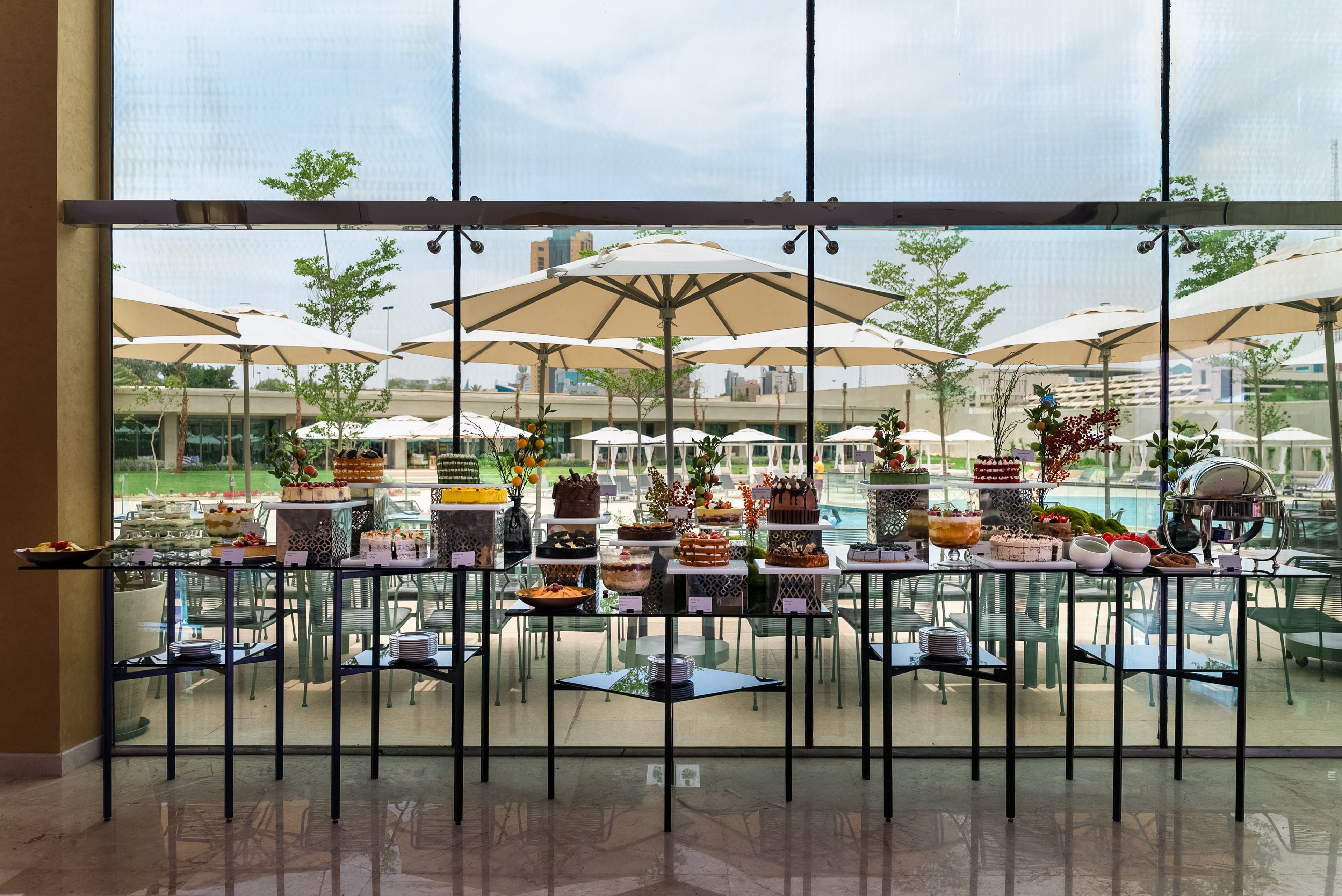 Zona de bufé con diversos postres y bebidas, mesas al aire libre con sombrillas, y vistas a una piscina y al horizonte urbano.