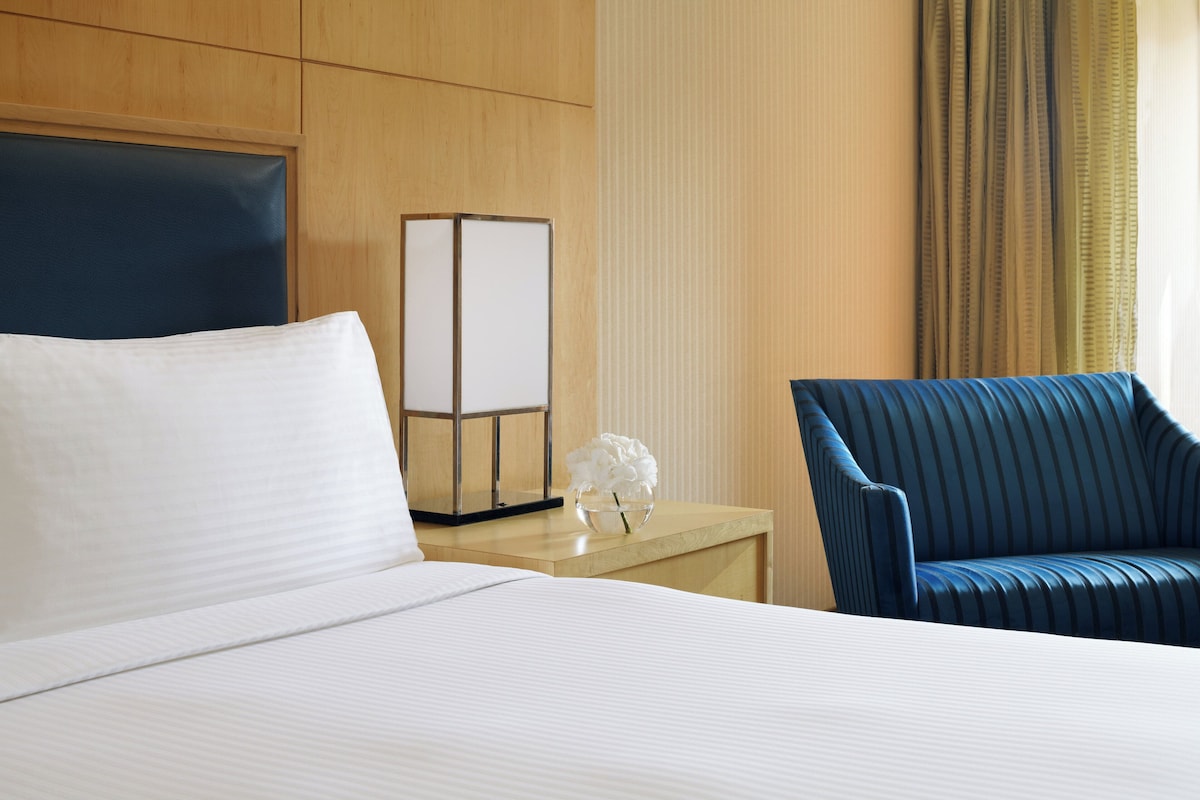 Una habitación de hotel con una cama, una mesita de noche, una lámpara, un jarrón con flores y una silla azul a rayas.