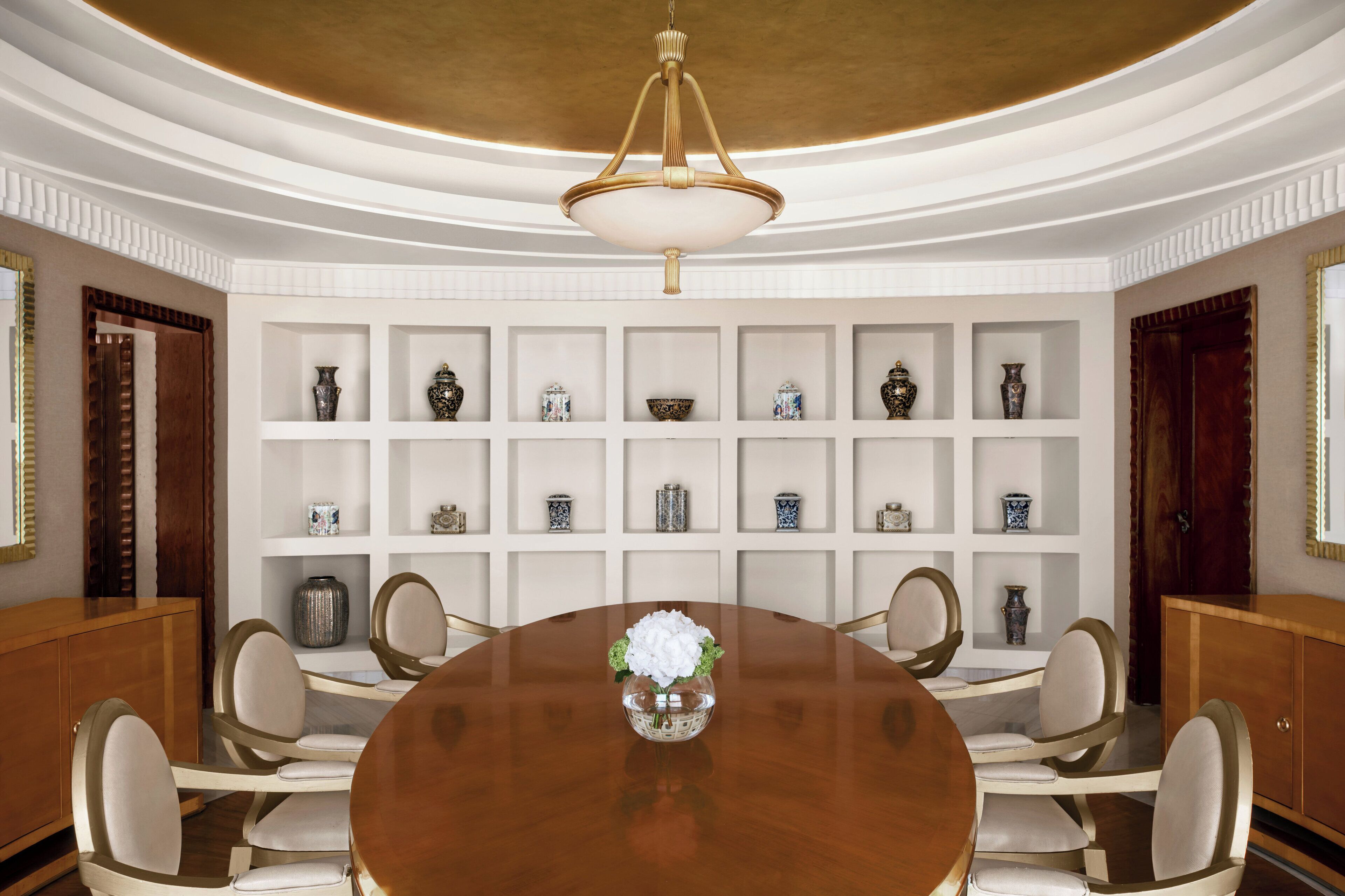 A formal dining room with a round table, six chairs, a chandelier, and a wall of shelves with decorative items.
