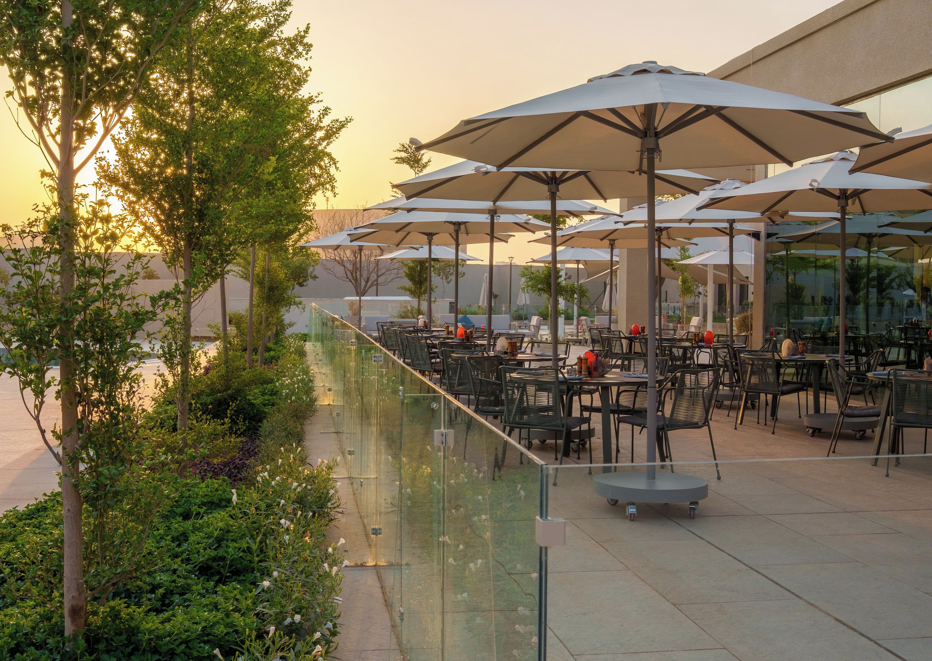 An outdoor patio area with tables and chairs, umbrellas, and a glass railing overlooking a garden.