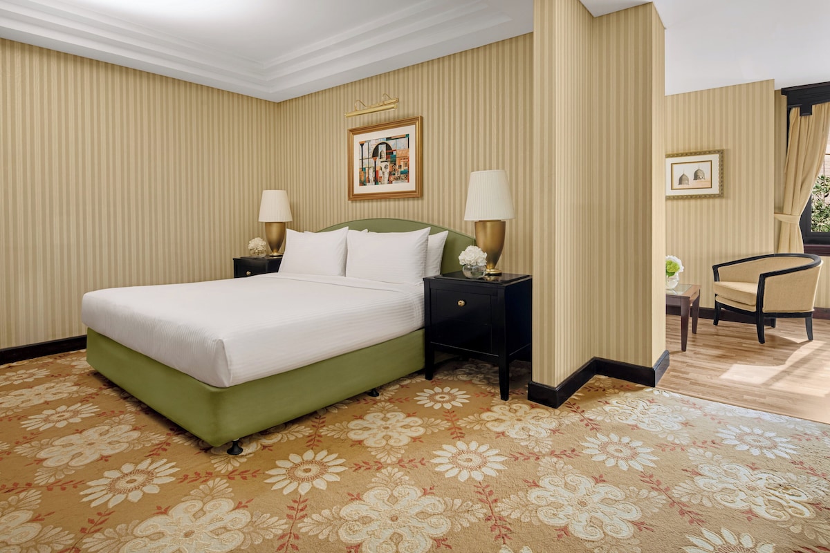 Habitación de hotel con una cama grande, dos mesitas de noche con lámparas, una silla y una pequeña mesa con un florero.