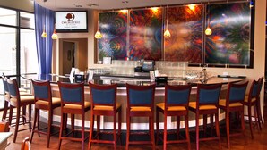 A bar area with a modern design, featuring a central bar counter, multiple bar stools, and large abstract wall art. - DoubleTree by Hilton Hotel Buffalo - Amherst (Buffalo)