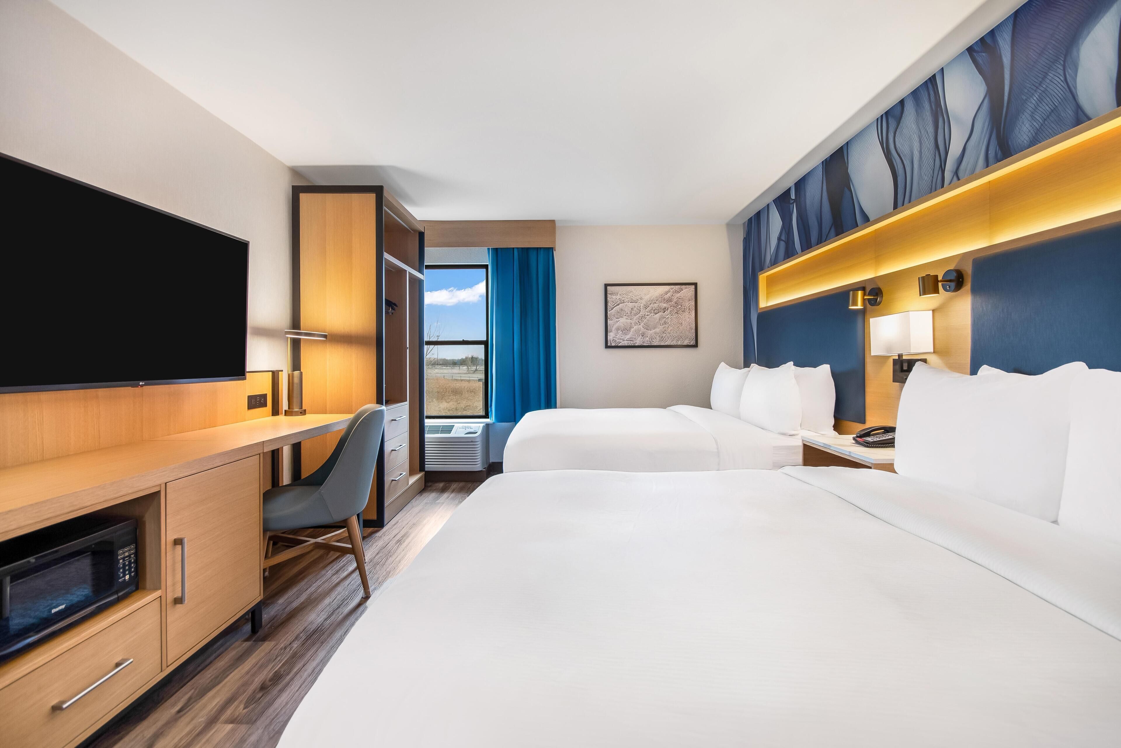 A hotel room with a bed, a desk, a chair, a flat-screen TV, a window with curtains, and a wall-mounted artwork.