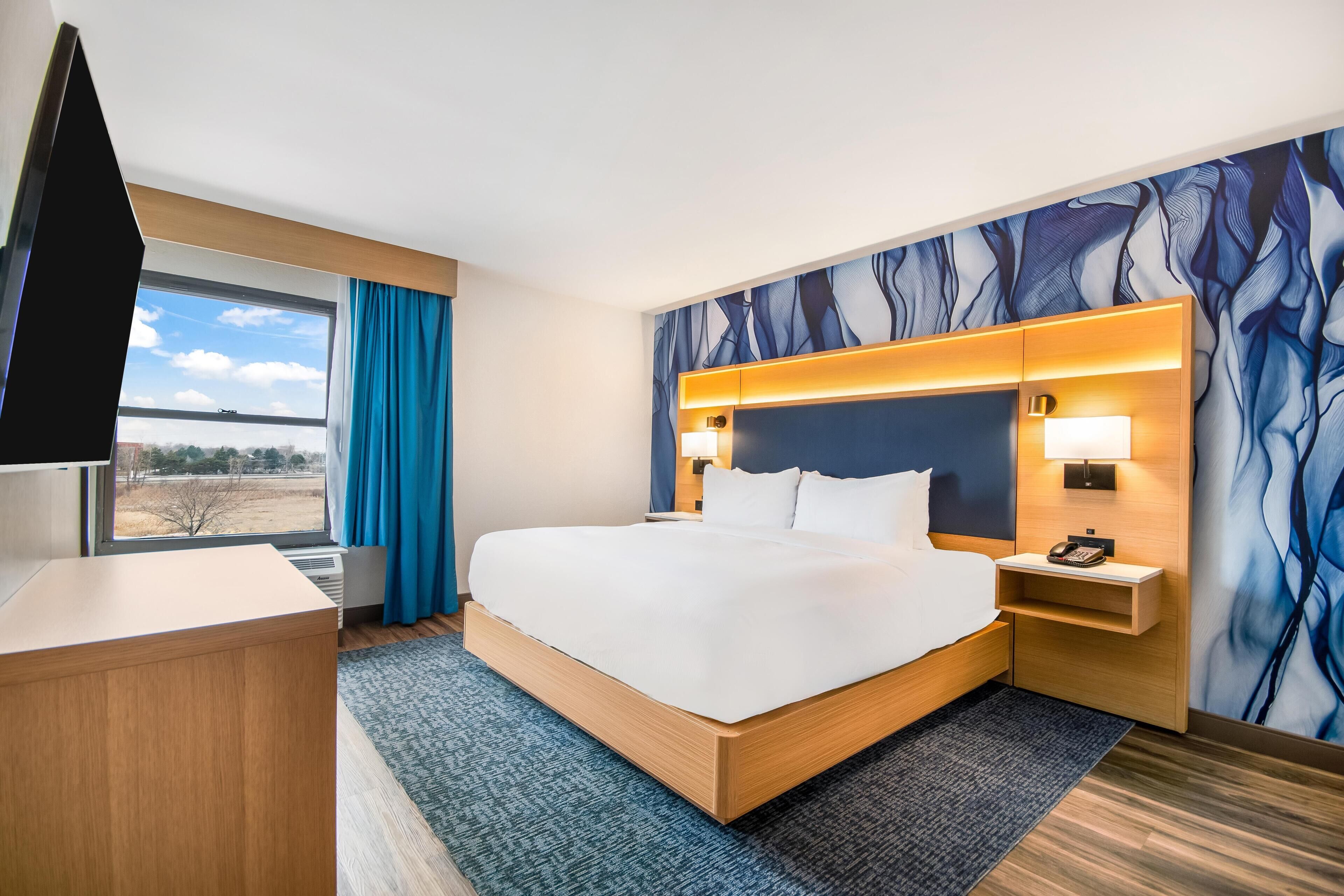 A hotel room with a bed, a desk, a TV, a window with curtains, and a wall with a blue abstract design.