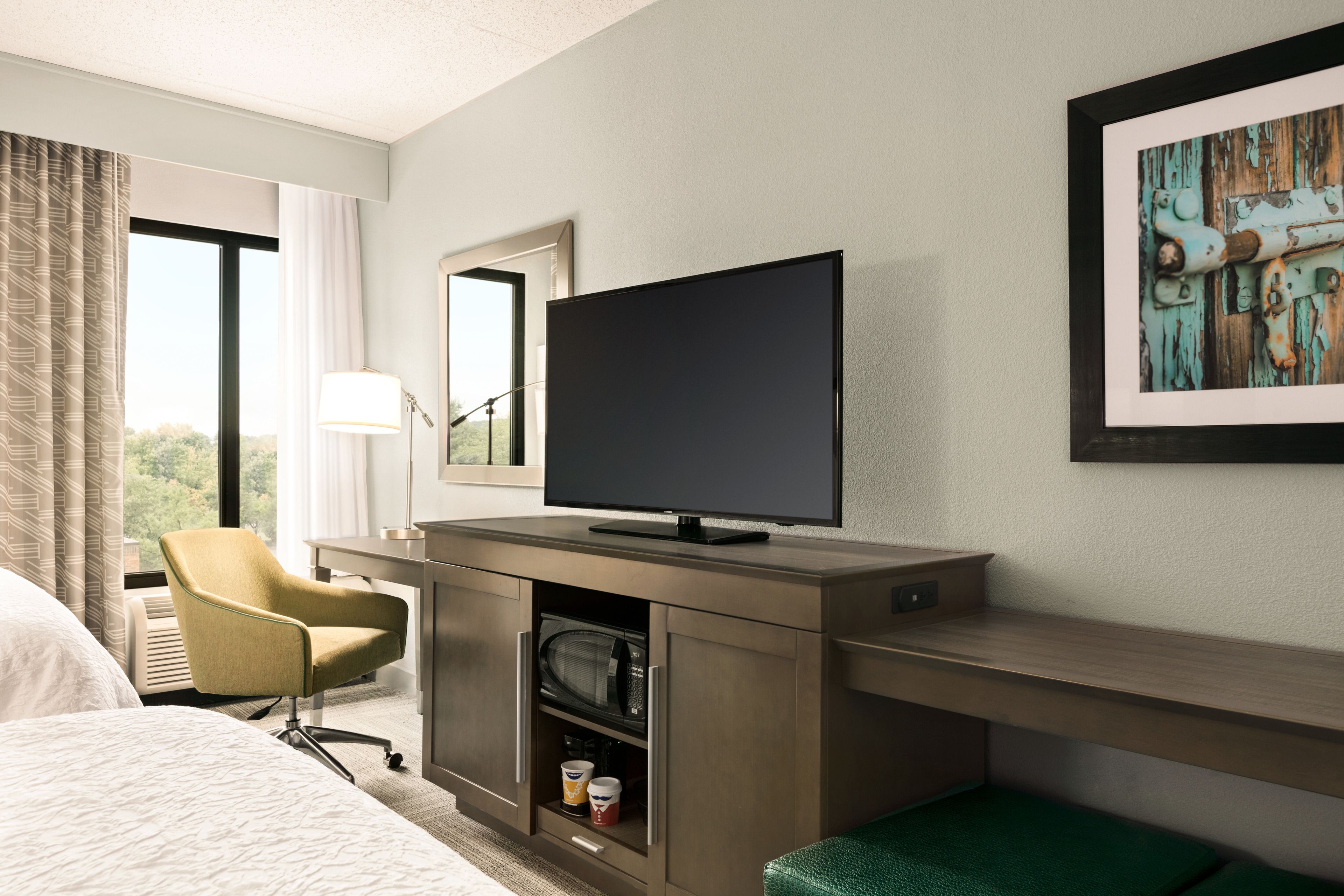 A hotel room with a bed, a desk with a television, a chair, a microwave, and a framed picture on the wall.