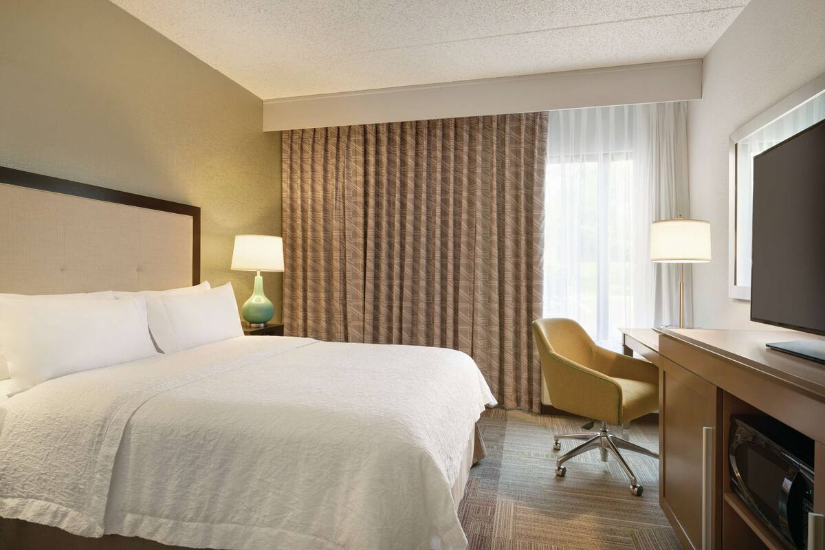 A hotel room with a large bed, a desk with a television, a chair, and a lamp.