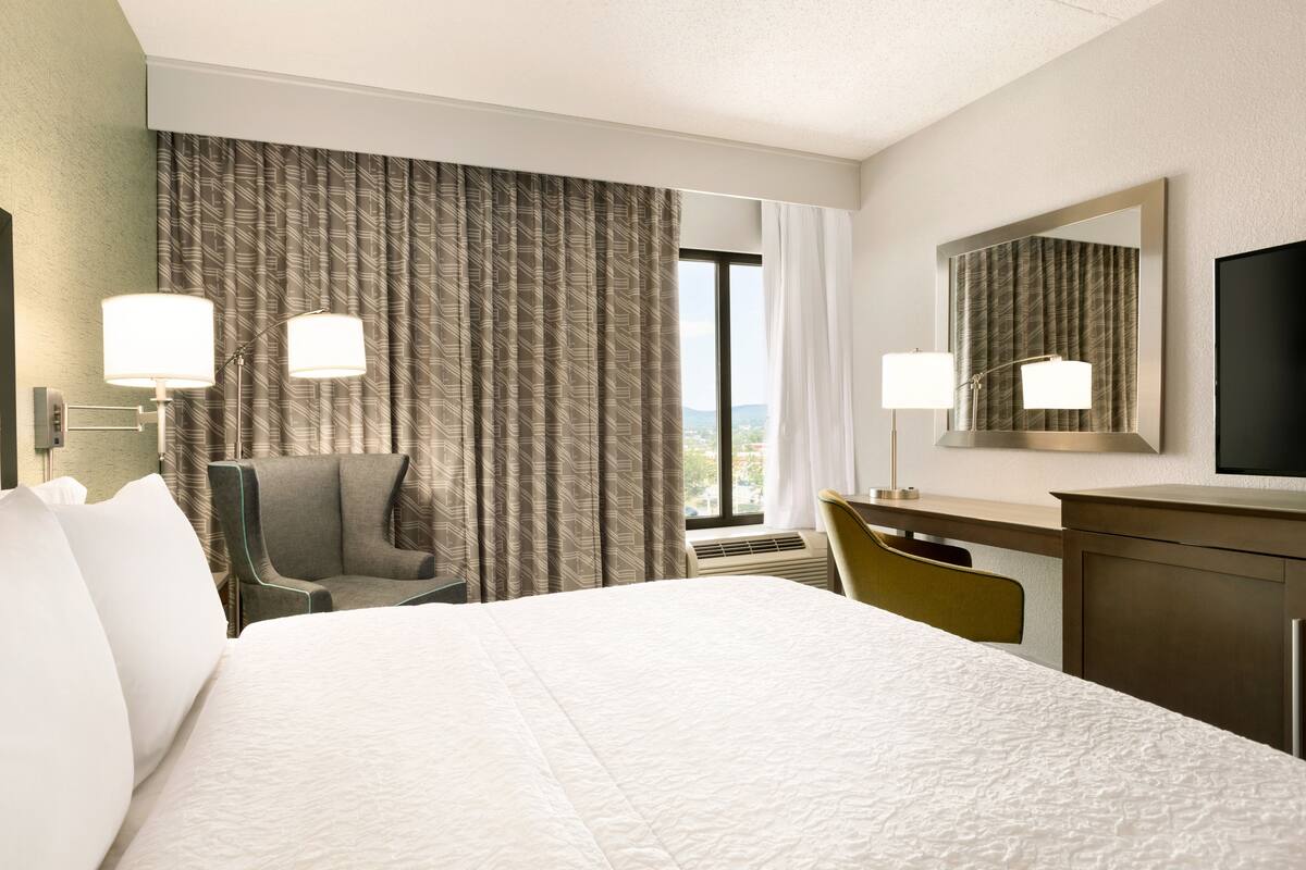 A hotel room with a large bed, a desk, a chair, a television, and a window with curtains.