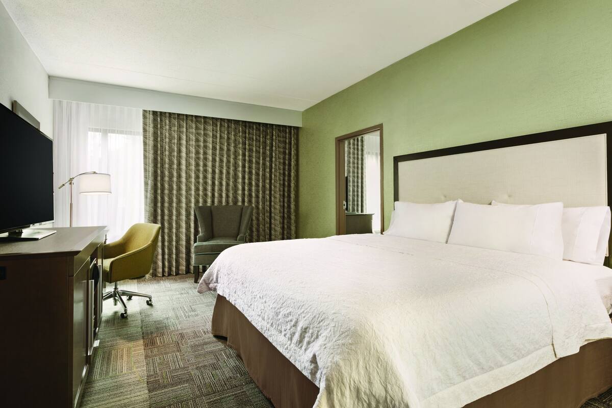 A hotel room with a large bed, a desk with a television, a chair, and a window with curtains.