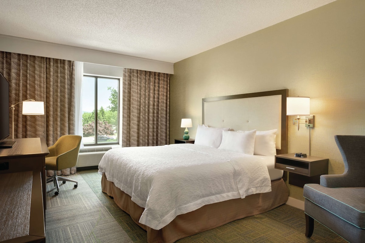 A hotel room with a large bed, a desk, a chair, and a window with curtains.
