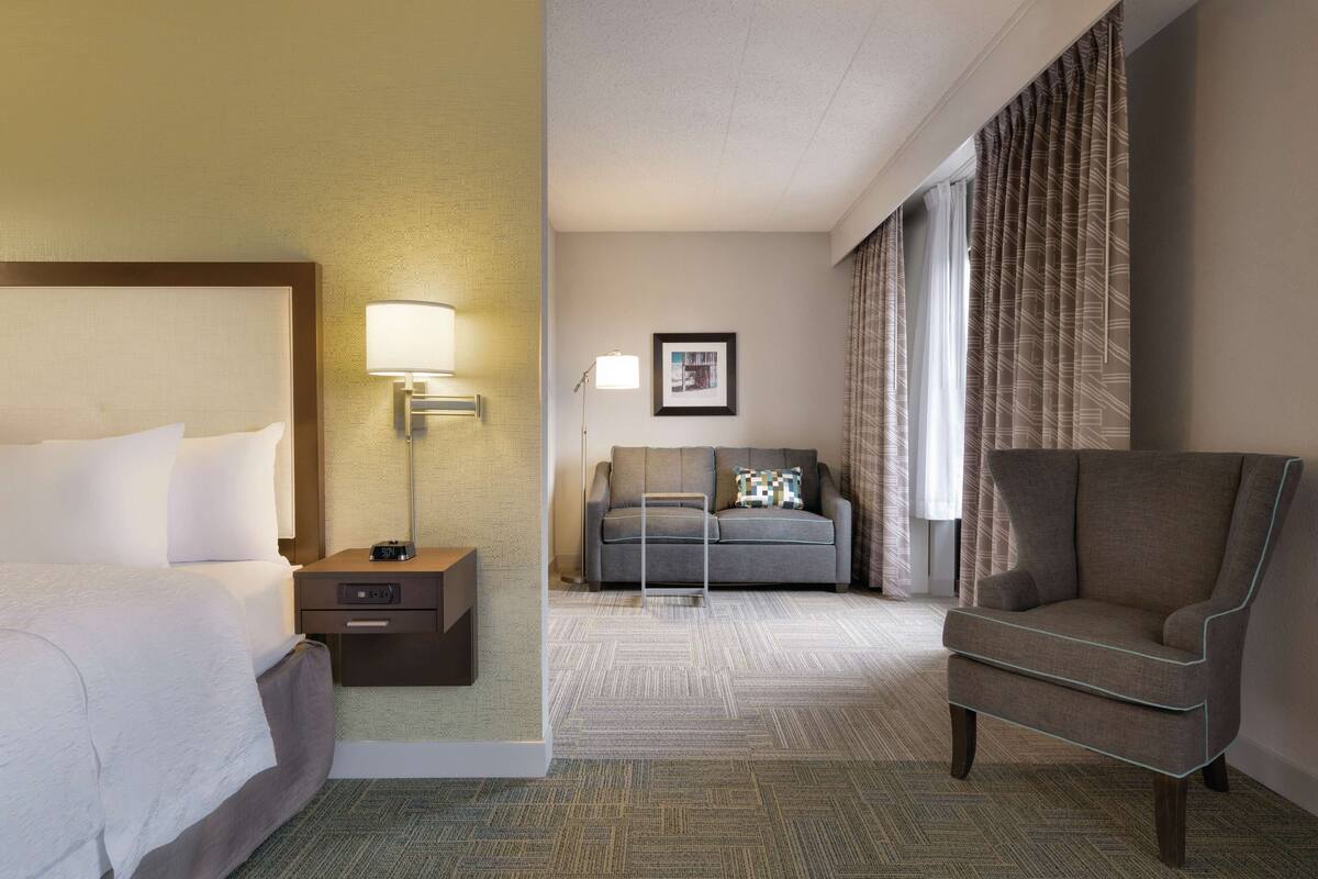A hotel room with a bed, a desk with a lamp, a sofa, a chair, and a large window with curtains.
