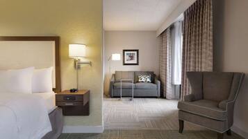 Habitación de hotel con cama, escritorio con lámpara, sofá, sillón y ventana grande con cortinas.