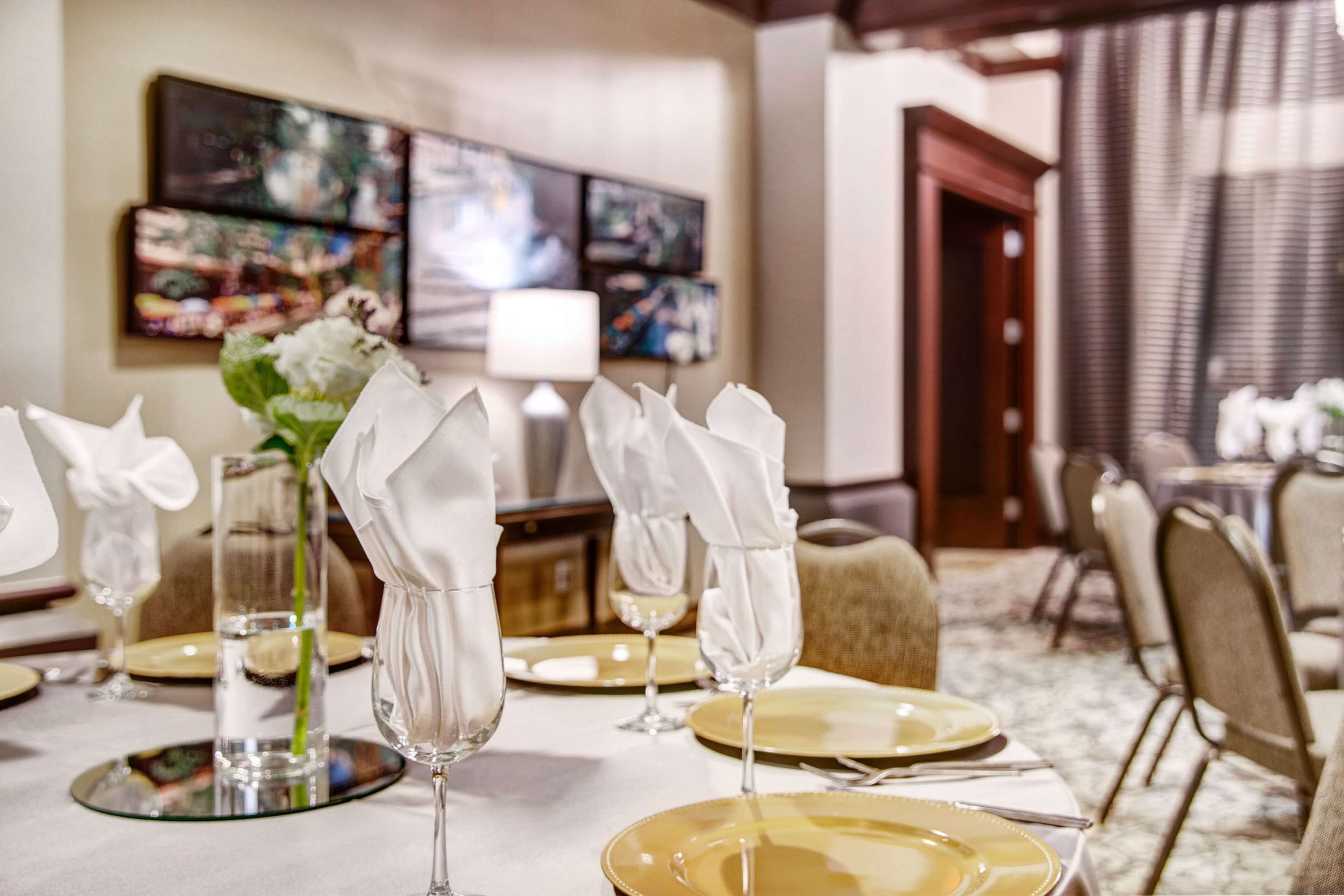 A dining table set with white napkins, clear glassware, and gold plates, with a vase of white flowers.