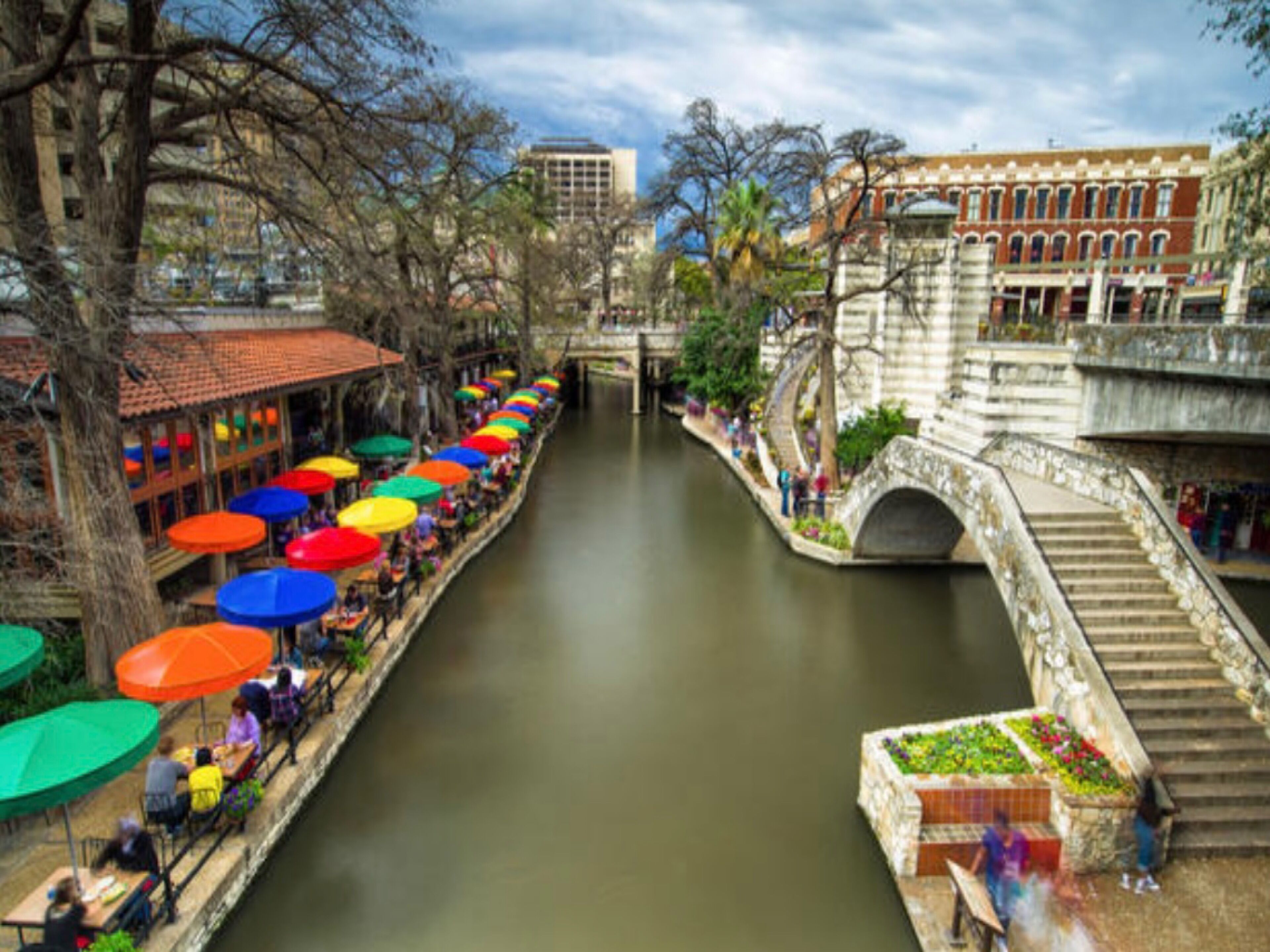 A canal with colorful umbrellas lining the sides, a stone bridge, and buildings in the background.