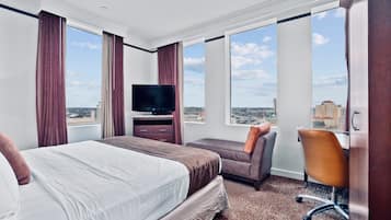 A hotel room with a large bed, a desk with a chair, a television, and a view of the city.