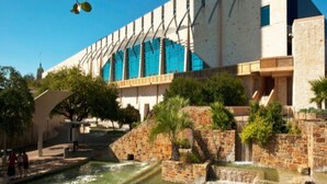 Un edificio moderno con fachada de vidrio extensa, un patio con fuentes de agua y un muro de piedra.