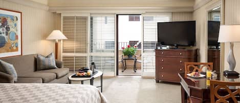 A hotel room with a bed, a sofa, a dining table with chairs, a TV, and a balcony with a table and chairs.
