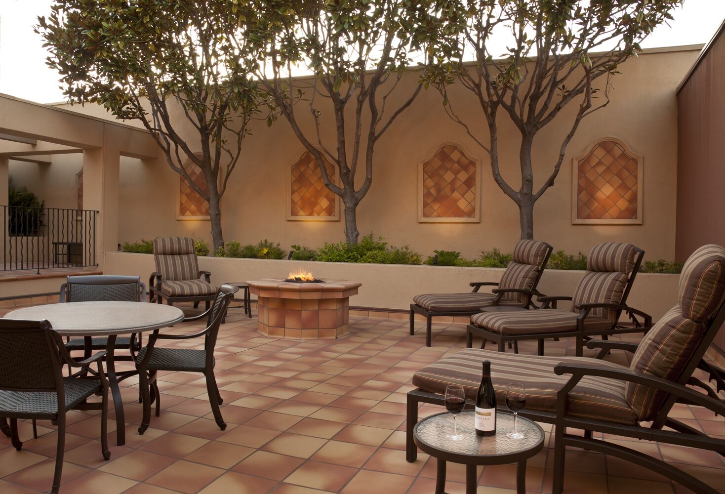 Une terrasse extérieure avec une table ronde, des chaises, un foyer et une bouteille de vin.