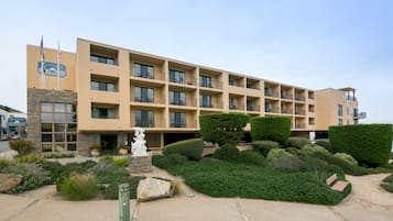 A multi-story hotel with a beige facade, balconies, and a landscaped front yard with shrubs and a statue.