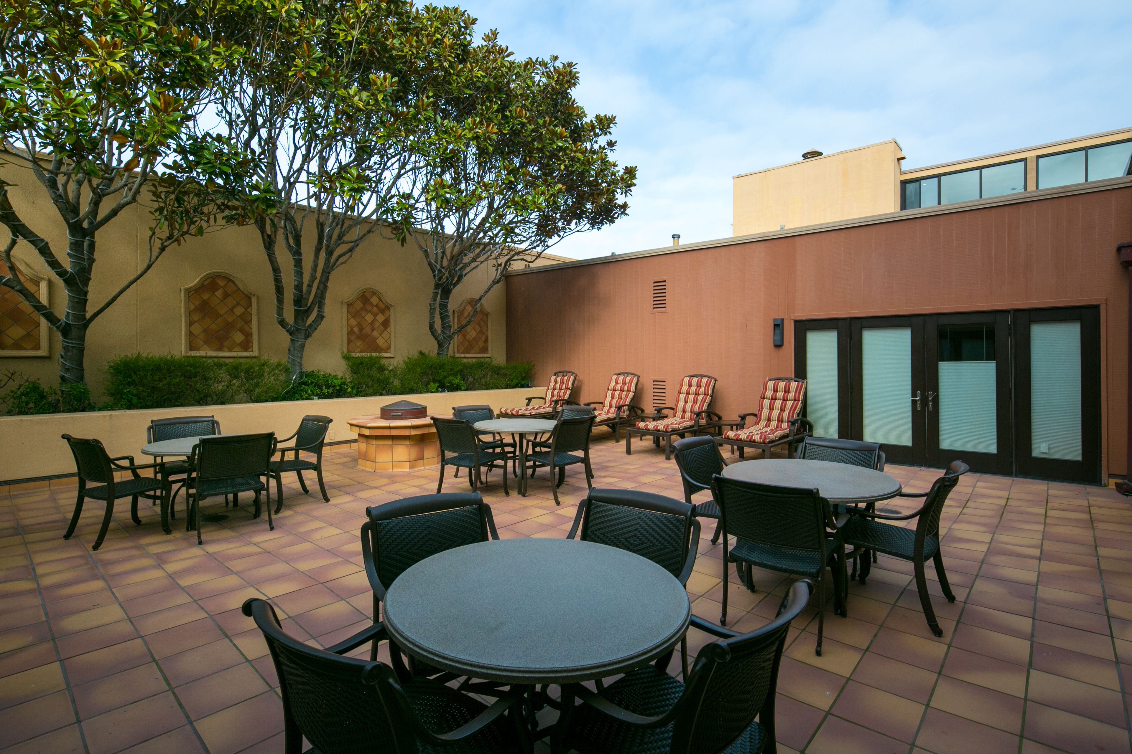 Un espace extérieur aménagé en terrasse, avec plusieurs tables et chaises, entouré de verdure et bordé d’un bâtiment doté de grandes fenêtres.