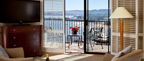 A living room with a TV, a wine bottle, and glasses on a table, a sofa, and a balcony with a view of the sea and mountains.