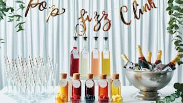 Disposición de bebidas con diferentes opciones, incluyendo champán y zumos de frutas, dispuestas sobre una mesa con fondo blanco y decoración de follaje.