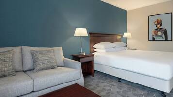 A hotel room with a grey sofa, a wooden nightstand with a lamp, a white bed, and a geometric-patterned carpet.