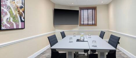 Un salón de conferencias con una larga mesa, sillas negras, una pantalla de proyector y una ventana con persianas.
