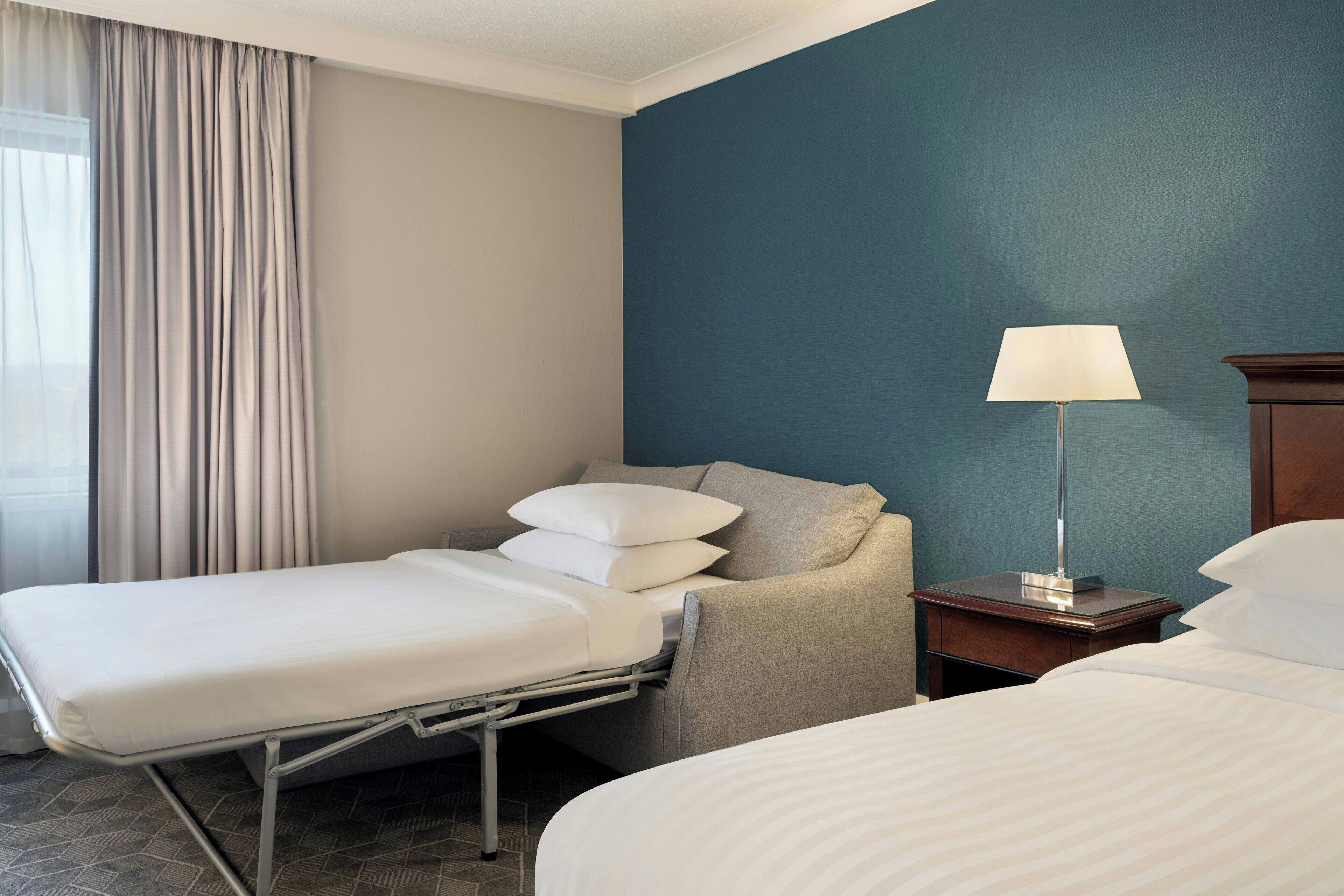 Camera d'albergo con due letti, un comodino con lampada, una sedia e una finestra con tende.