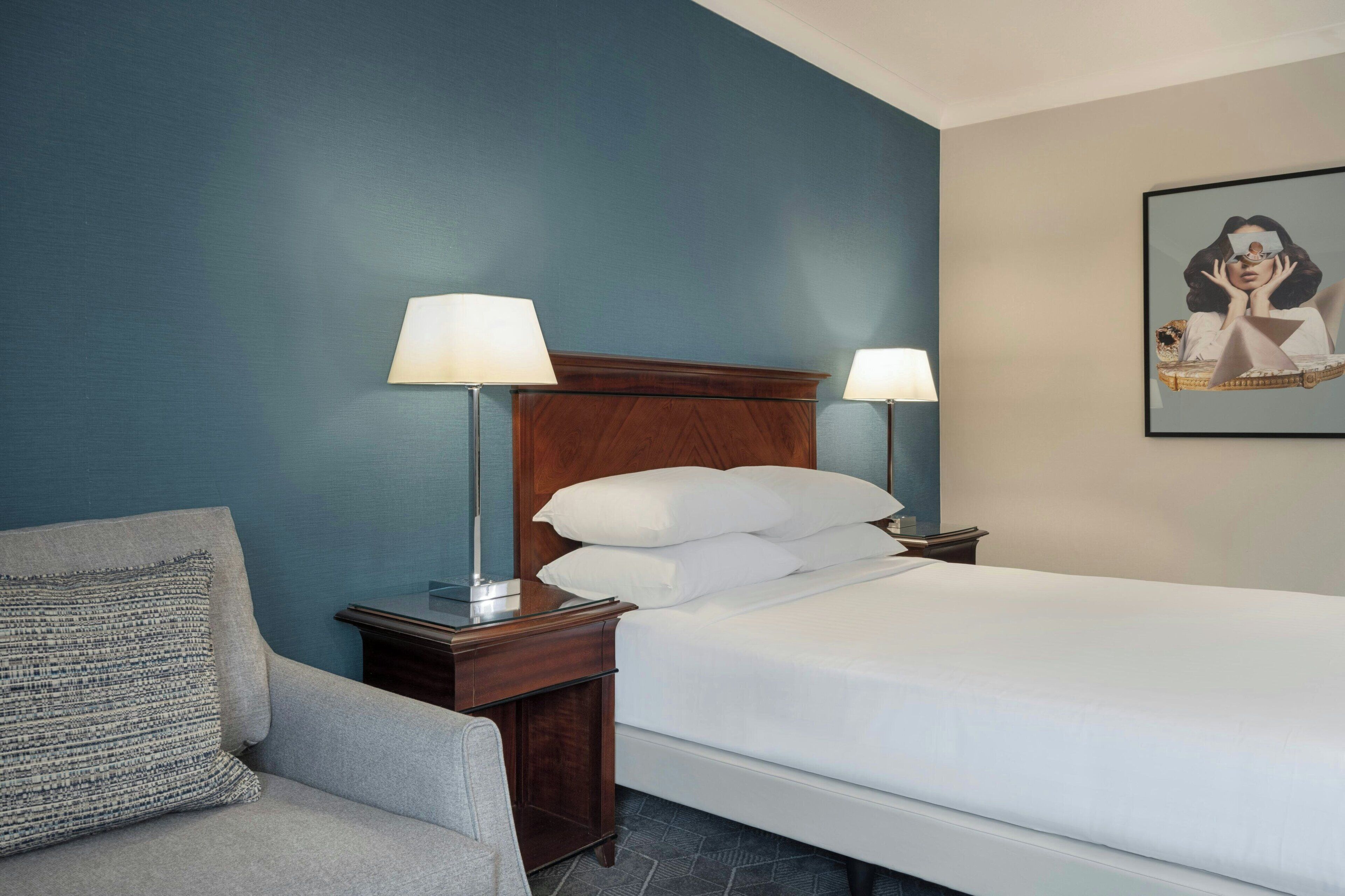 A hotel room with a bed, two bedside tables with lamps, a chair, and a wall-mounted artwork.