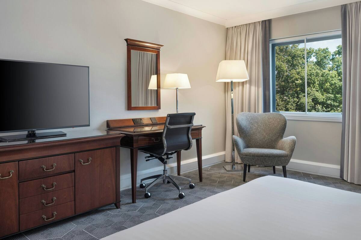 Une chambre d’hôtel comprenant un lit, un bureau avec une chaise, une télévision, une lampe et une fenêtre avec des rideaux.