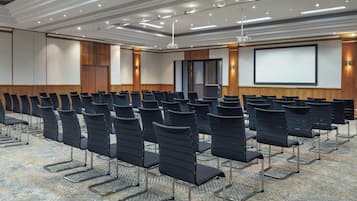 Uma sala de conferências com fileiras de cadeiras pretas, uma tela grande e piso coberto de carpete.