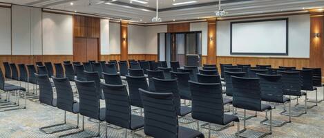 Uma sala de conferências com fileiras de cadeiras pretas, uma tela grande e piso coberto de carpete.