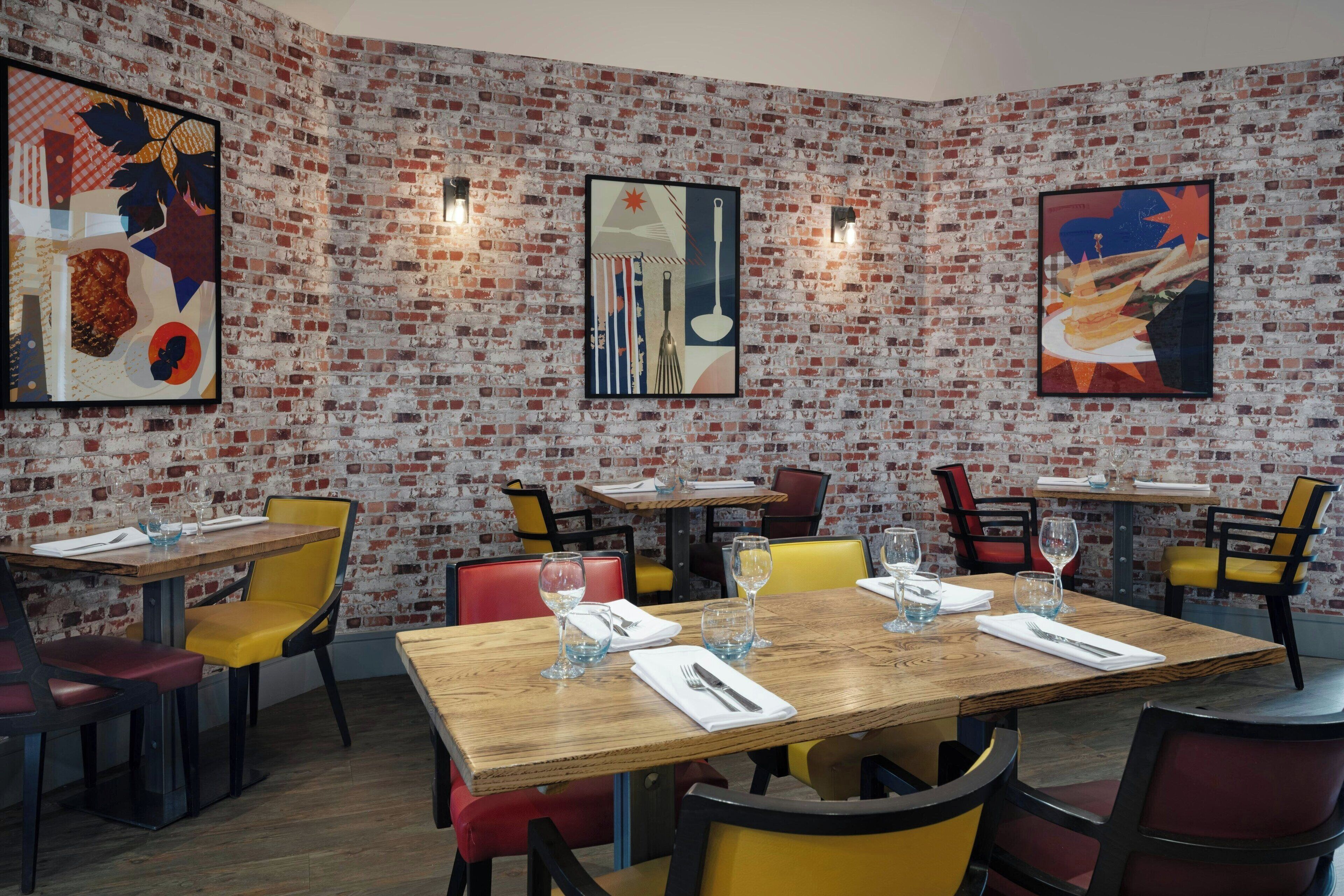 A restaurant with brick walls and colorful chairs. Tables are set with glasses and cutlery.