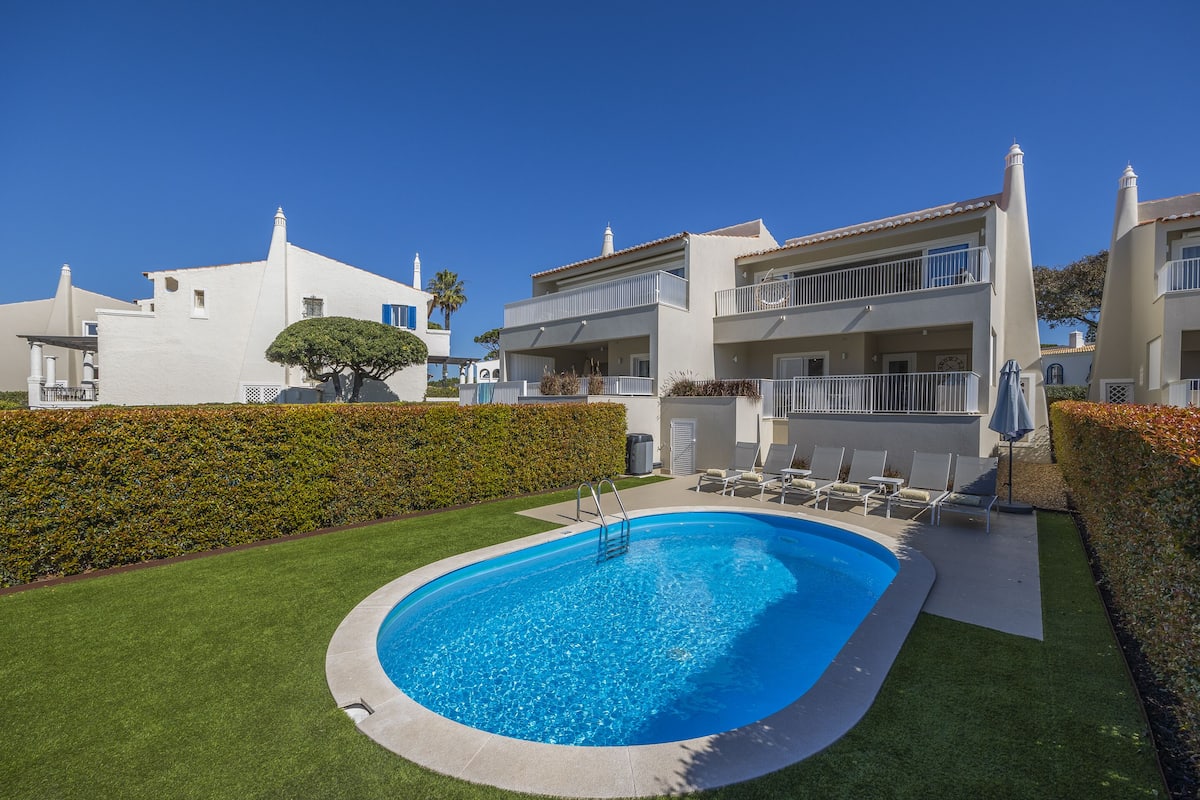 A swimming pool surrounded by a well-maintained lawn, with a building featuring balconies and a clear blue sky in the background.