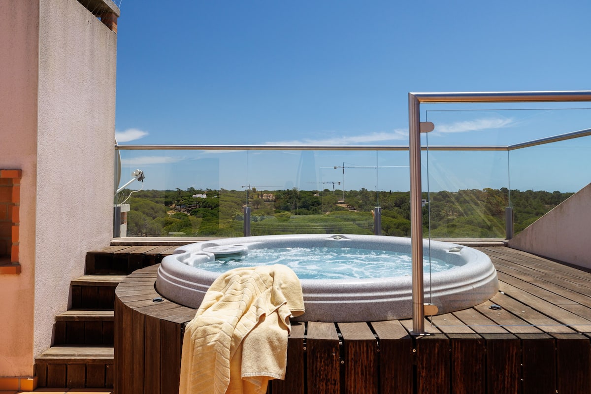 A hot tub on a wooden deck with a glass railing, overlooking a green landscape.