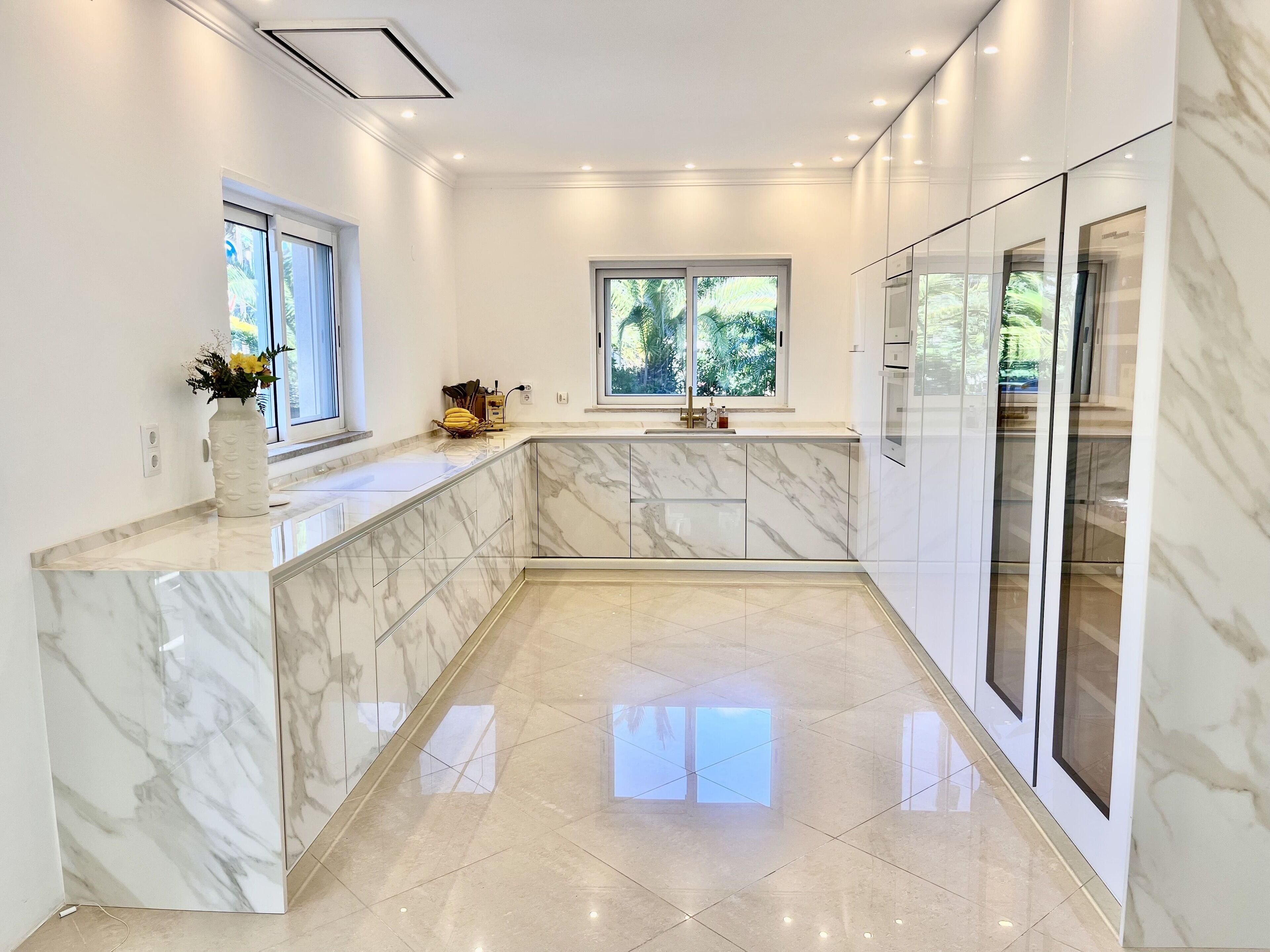 A modern kitchen with marble countertops, a built-in oven, and a window providing a view of greenery.