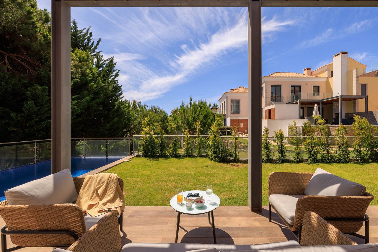 Une terrasse extérieure aménagée avec des meubles en osier, une petite table avec des boissons et des collations, et une vue sur une piscine et des immeubles résidentiels.