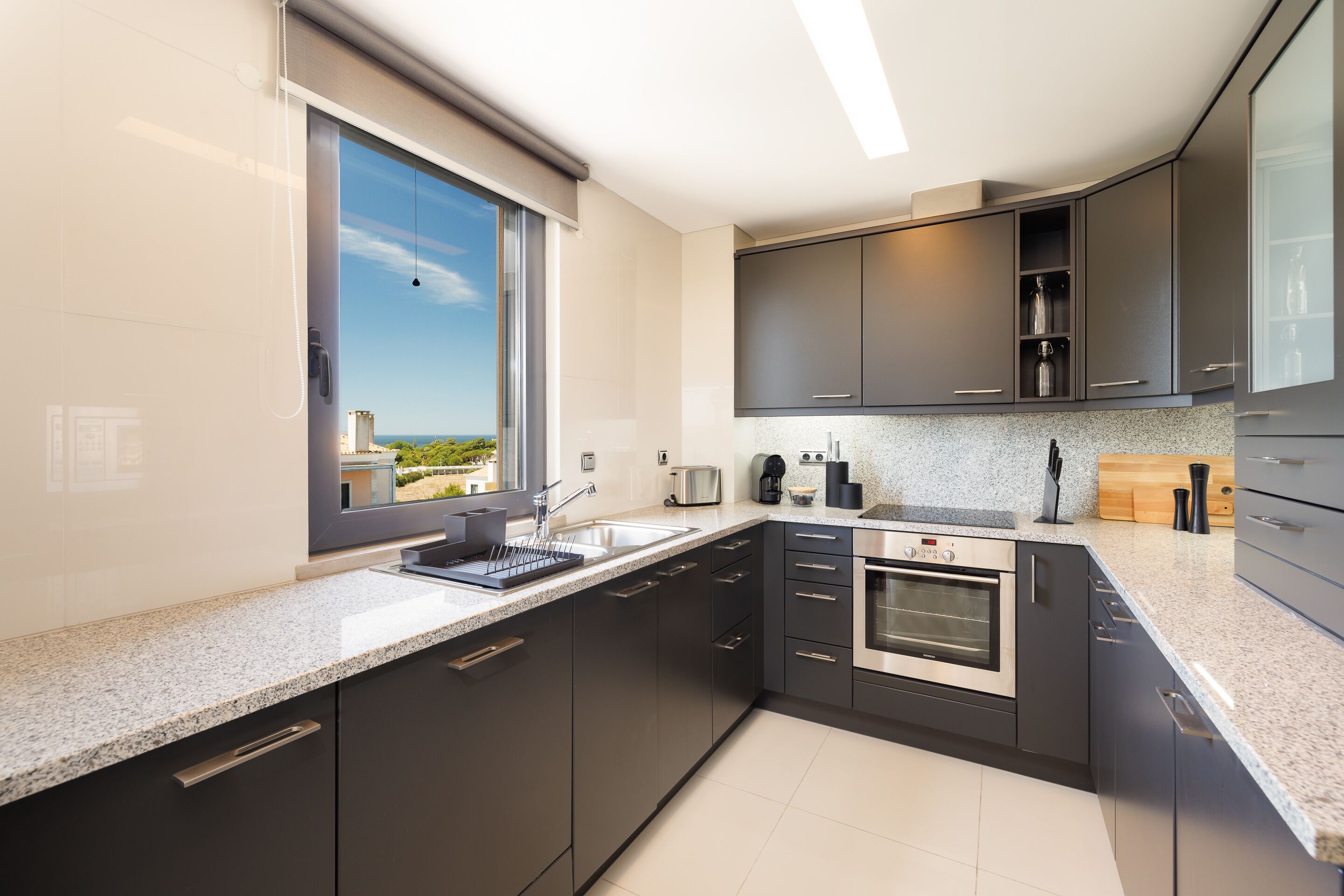 A modern kitchen with dark cabinets, a stainless steel oven, a built-in coffee maker, and a window offering a view of greenery.