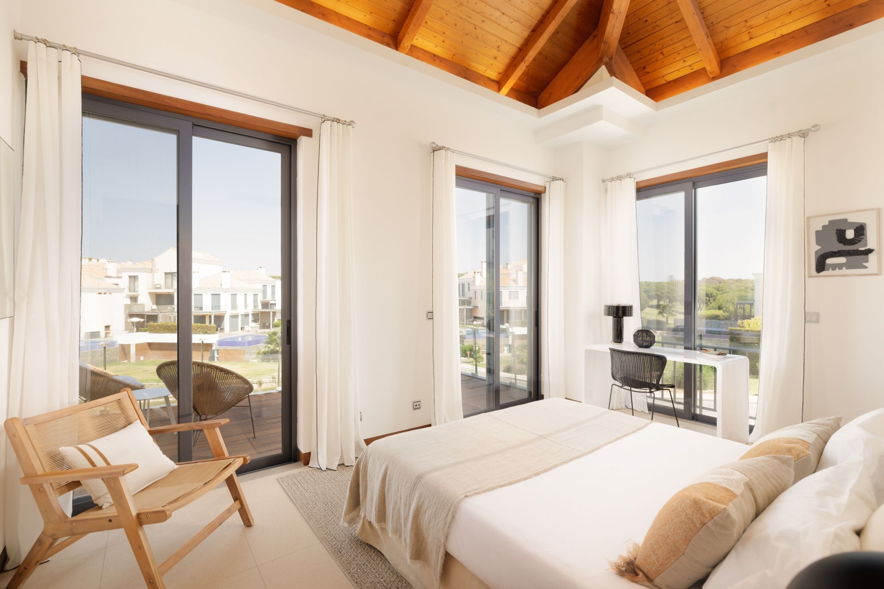 Ein Schlafzimmer mit einem großen Bett, einem Holzstuhl und einem Schreibtisch mit Stuhl. Es gibt große Fenster und einen Balkon mit Blick auf Gebäude.