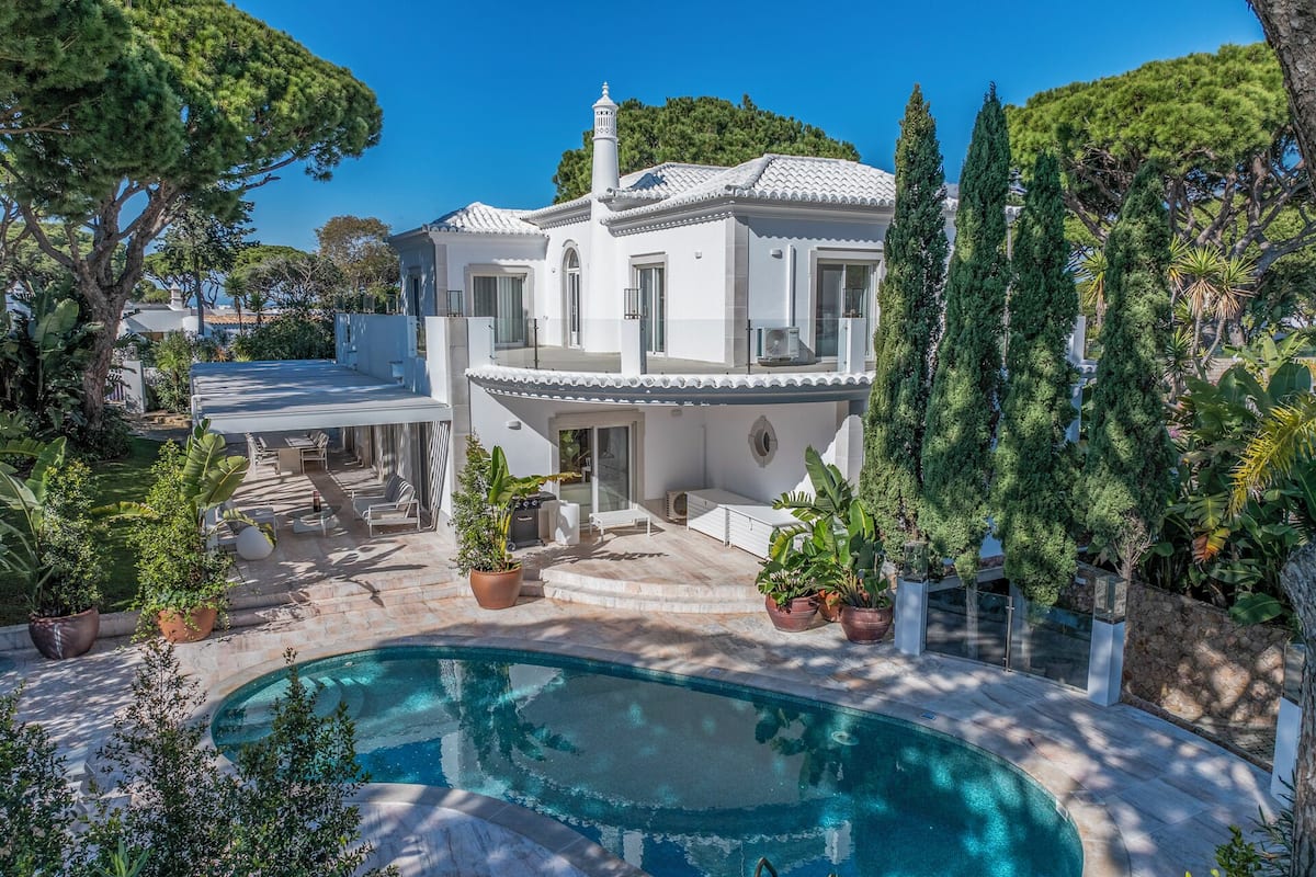 A two-story house with a swimming pool, surrounded by lush greenery and a clear blue sky.